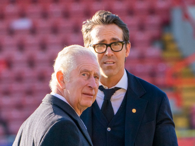 Prince Charles looking behind himself looking at the camera standing in front of Ryan Reynolds, who looks confused
