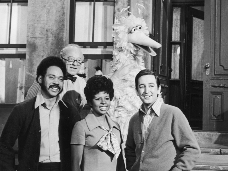black and white photo of several adult actors in typical 1960s fashion smiling and posing with Big Bird on the set of Sesame Street