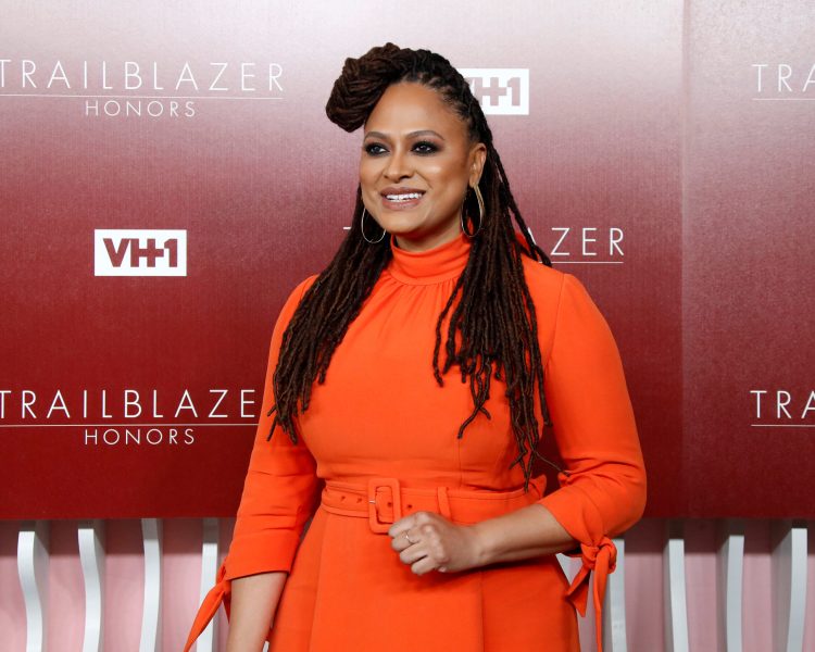 Ava DuVernay smiles in orange dress against dark red backdrop