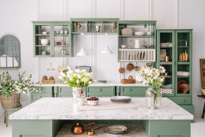 A beautiful kitchen with green cabinets and a marble countertop with flowers and open shelving