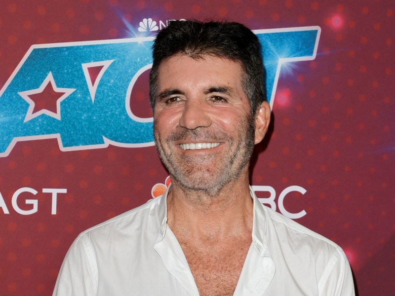 Simon Cowell smiles in white top against red backdrop