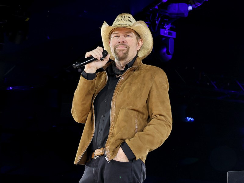 Toby Keith singing into a microphone on stage in a tan coat and cowboy hat