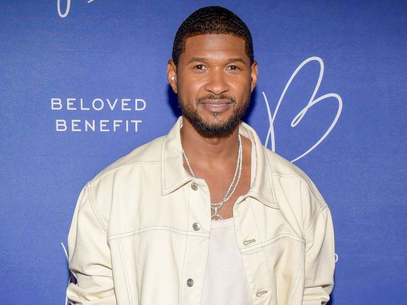 Usher smiles in white shirt and matching jacket against blue backdrop