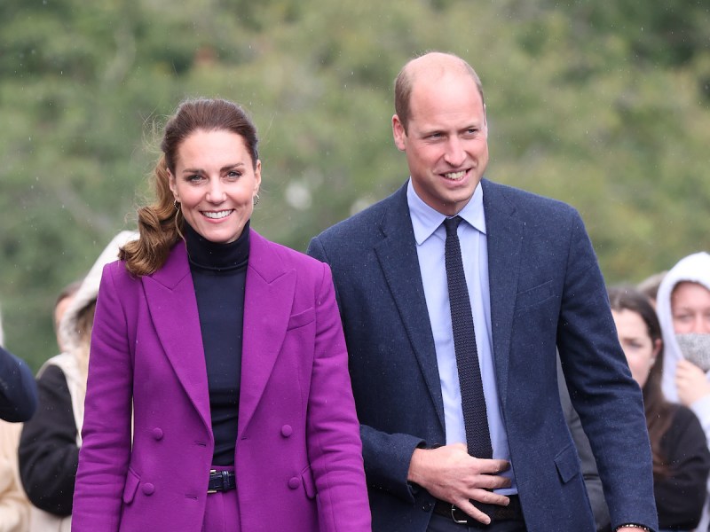 Kate Middleton smiling in a purple pantsuit with Prince William in a navy suit