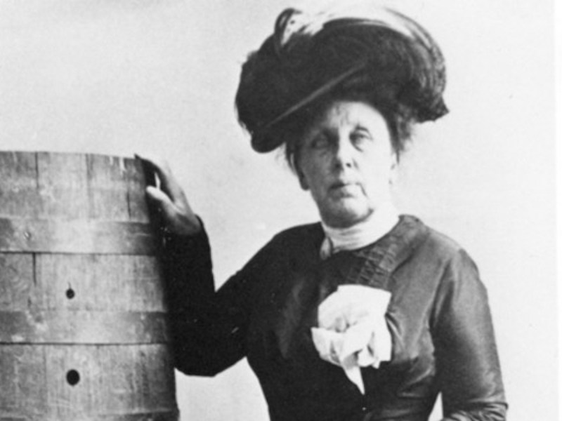 cropped headshot from a black and white photo of a mature looking Annie Taylor in dress and hat standing next to the barrel she went over the falls in