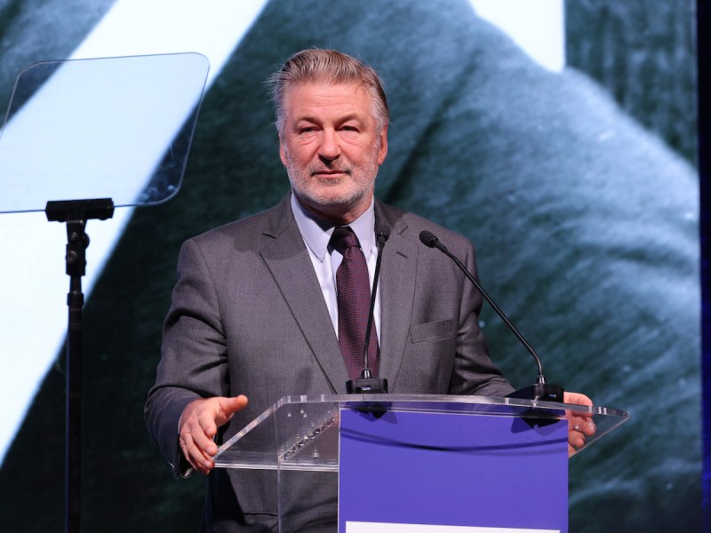 Alec Baldwin in a gray suit giving a speech at a podium on stage