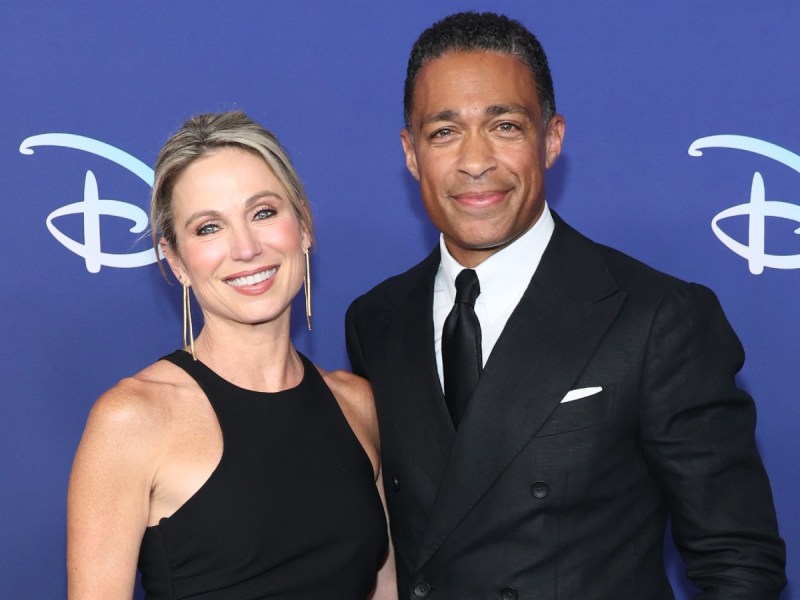 Amy Robach (L) in black dress posing next to TJ Holmes, who is wearing a black suit and tie