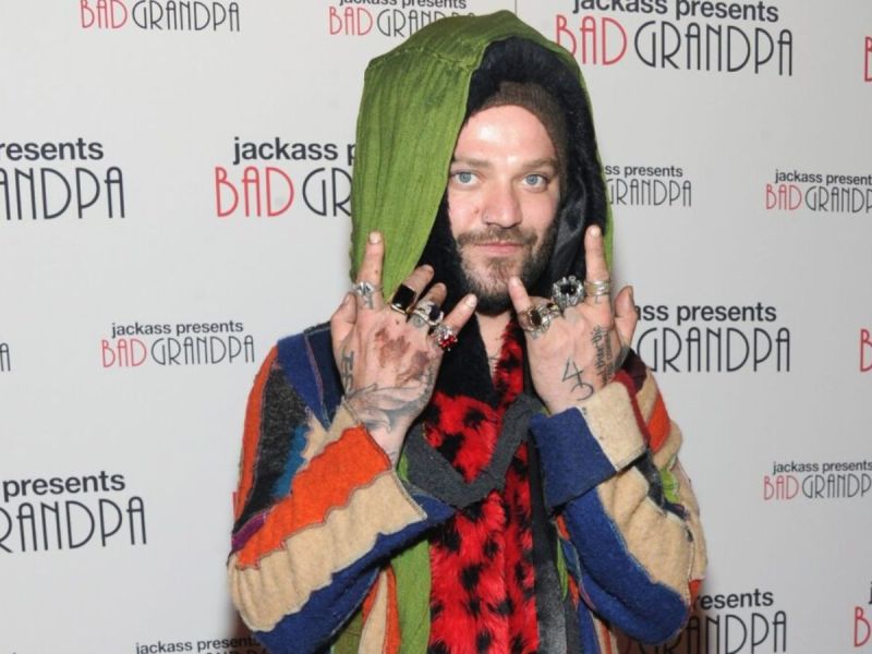 Bam Margera wearing a multicolored sweater with his hood up, holding up the "rock on" symbol.