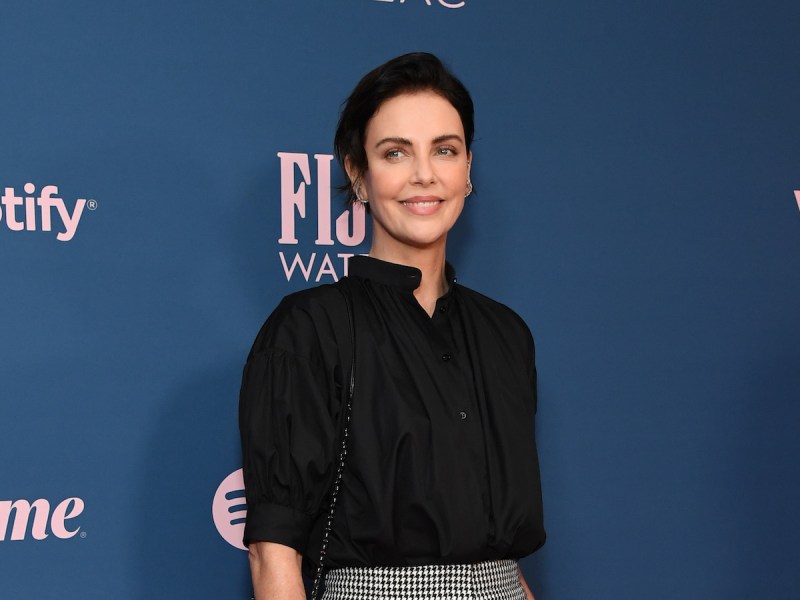 Charlize Theron smiling in a black blouse and grey skirt with short black hair