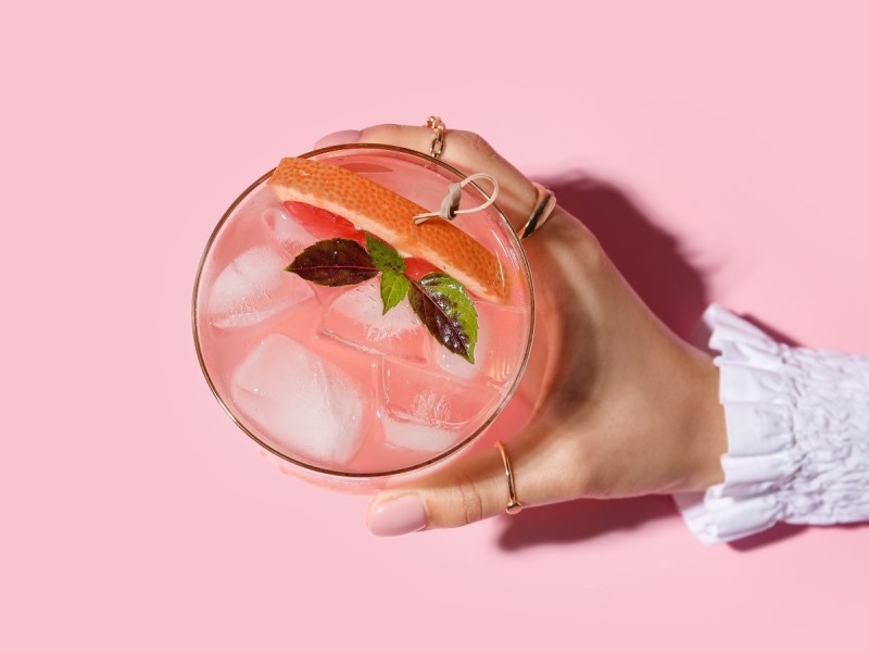 Overhead view of a woman's right hand holding a pink cocktail a pink surface
