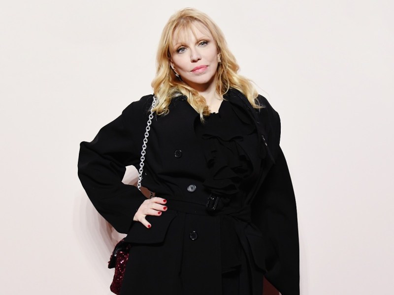 Courtney Love standing in black jacket while holding black crossbody purse against off-white backdrop