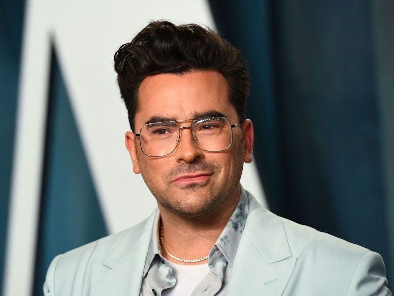 close up of Dan Levy smiling in a light blue shirt with large thin-rimmed glasses