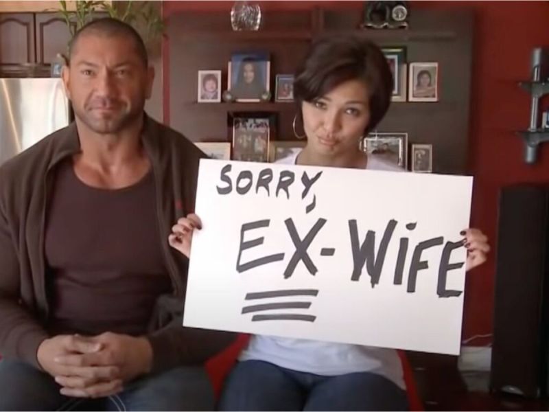 David Bautista sits next to his ex-wife Angie, who is holding up a sign that says "Sorry, ex-wife"