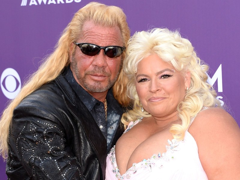 Dog the Bounty Hunter (L) in leather jacket posing with Beth Chapman, who is wearing a white top against purple backdrop