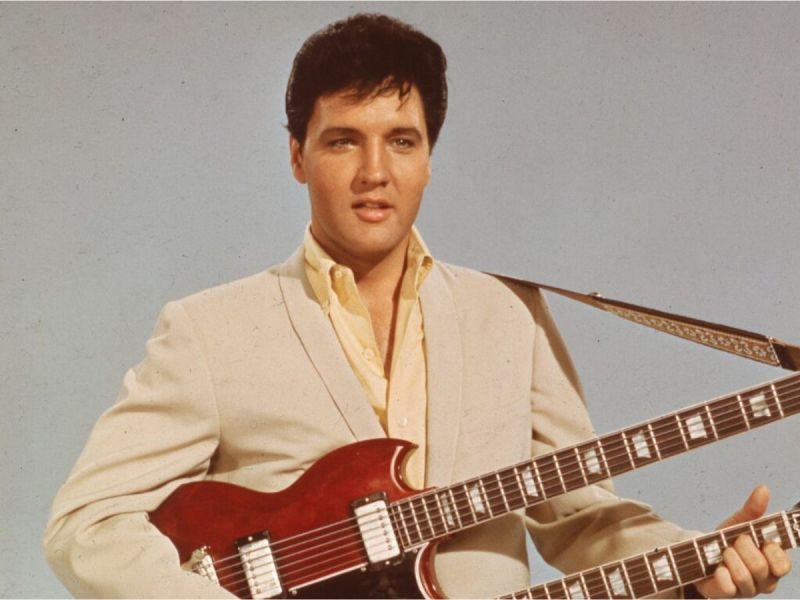 Elvis Presley smiles while holding a red guitar