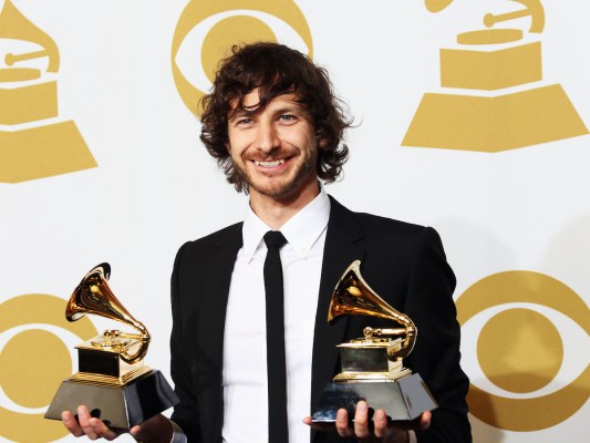 Gotye holding up two grammys in a black suit and tie