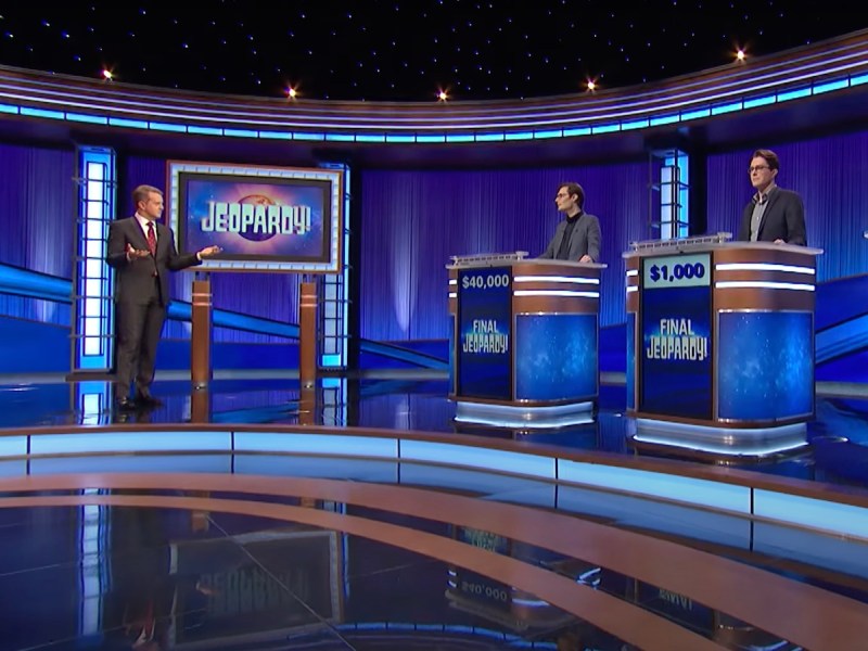 screenshot of a wide shot of the Jeopardy! stage with Ken Jennings speaking to contestants at their podiums