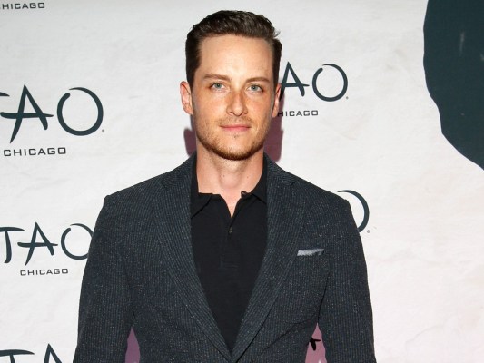 Jesse Lee Soffer smiles in black top and charcoal blazer against white backdrop