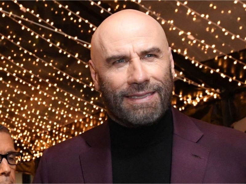 A close up of John Travolta in a black turtleneck and maroon jacket