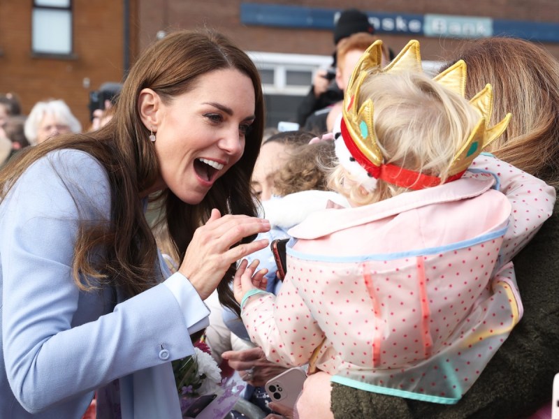 Kate Middleton (L) in powder blue jacket laughing with baby, who is wearing a toy crown