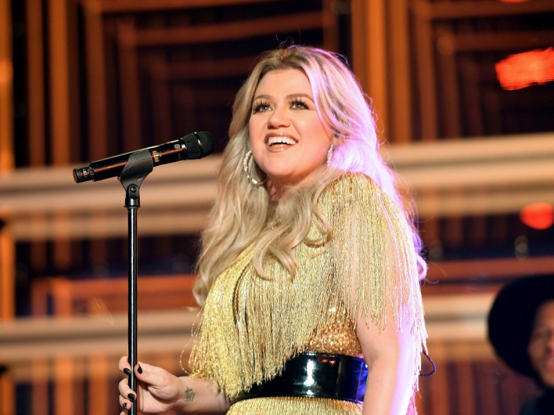 Kelly Clarkson smiles in gold outfit standing in front of microphone