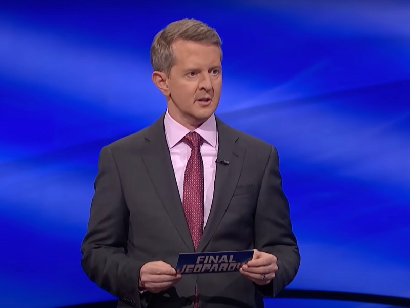 screenshot of Ken Jennings hosting Jeopardy! in a gray suit with his mouth slightly open