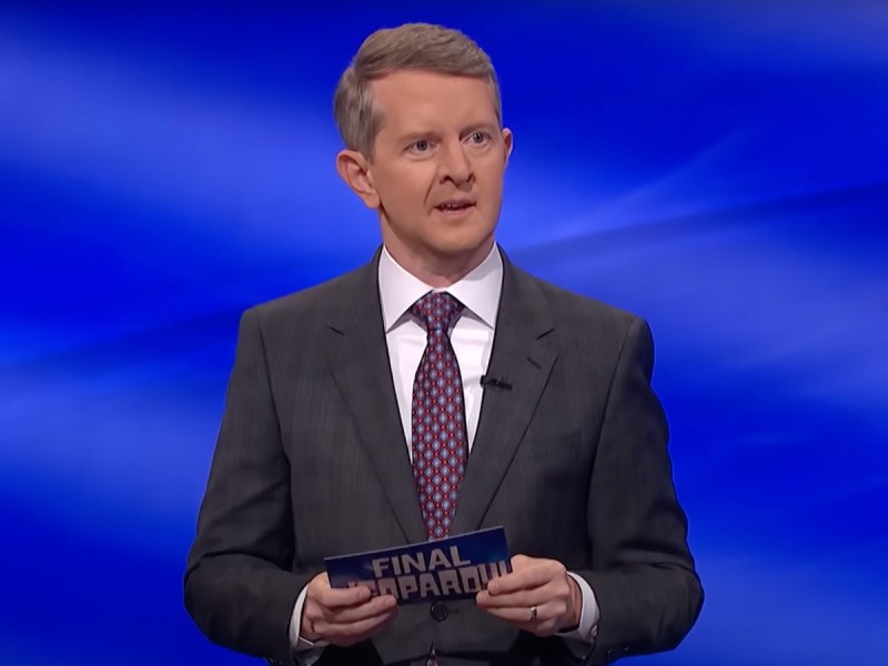 Ken Jennings holds cue card on 'Jeopardy!' stage while wearing a suit with red and blue patterned necktie