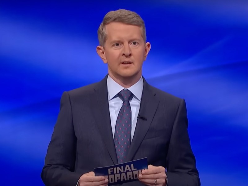 Ken Jennings wearing suit on 'Jeopardy!' stage speaking and holding cue card