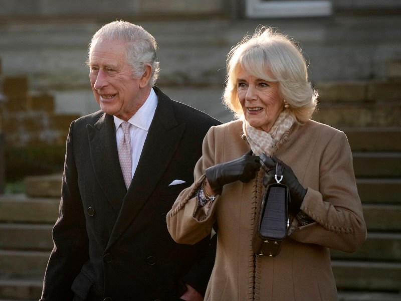 King Charles in a black suit and coat walking with Queen Consort Camilla in a tan coat
