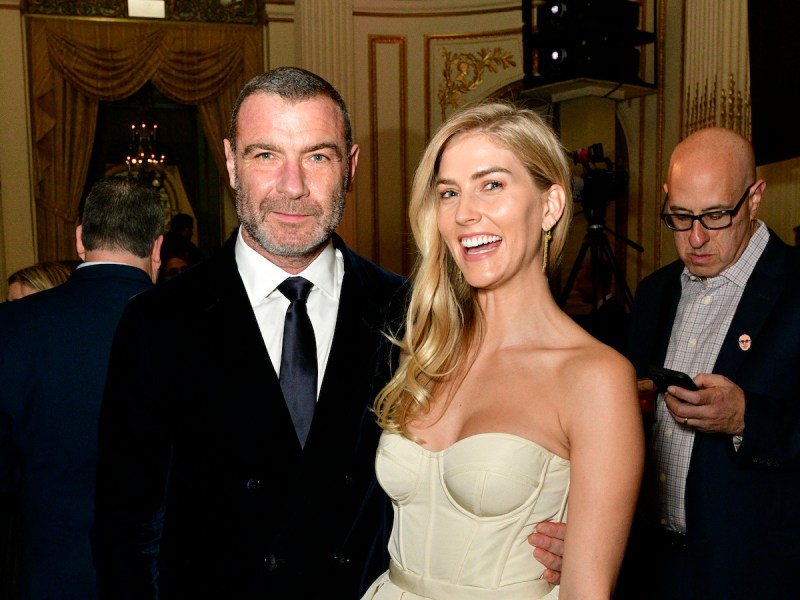 Liev Schreiber in a black suit smiling with his arm around a laughing Taylor Neisen in an eggshell colored dress