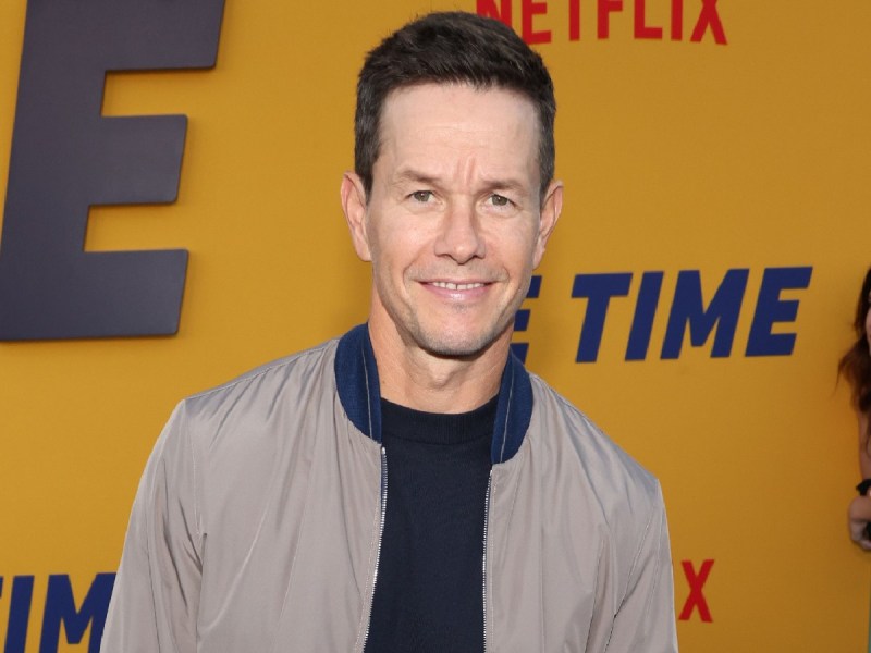Mark Wahlberg smiles in navy blue top and beige jacket against yellow backdrop