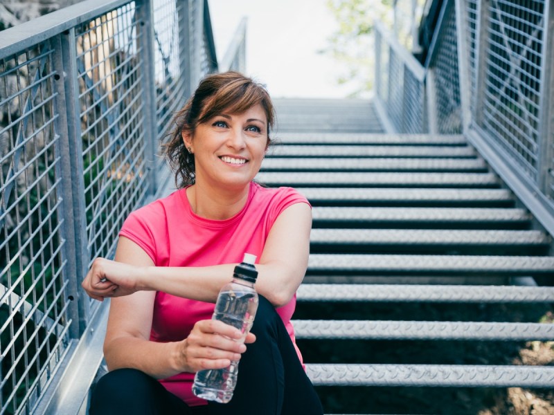 Middle-aged woman sits on steps holding water bottle and wearing pink exercise clothing