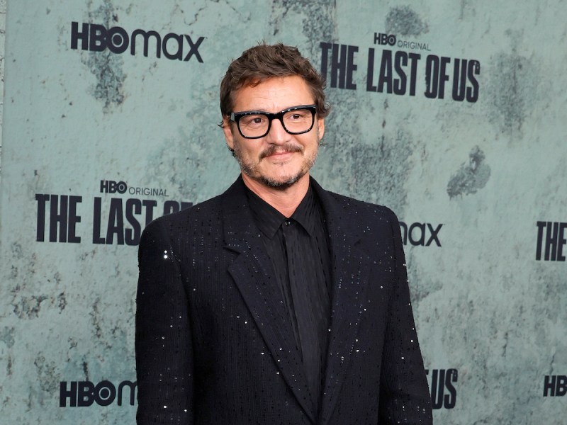 Pedro Pascal smiling in a suave black jacket and suit with black rimmed glasses