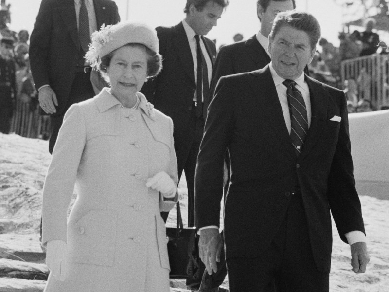 Queen Elizabeth II and Ronald Reagan walking in front of several men in suits, both looking at the camera
