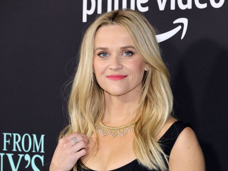 Reese Witherspoon smiling in a black dress with a silver ring and gold necklace
