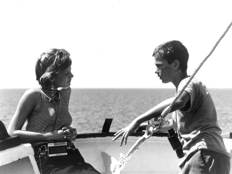 Teenaged Martha Plimpton leaning on a boat's rail while teenaged River Phoenix hangs his arms over a rope and speaks to her