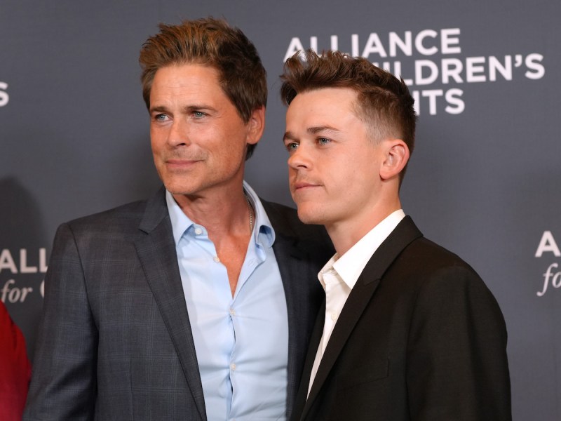 Rob Lowe in a grey suit smiling with son John Owen in a black suit