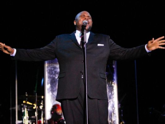 Ruben Studdard performing and wearing a black suit with his hands in the air.