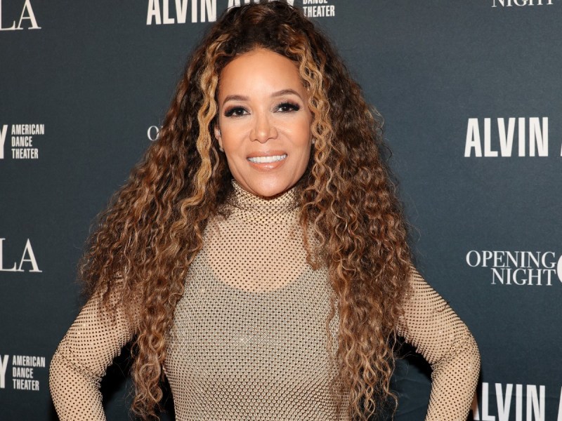 Sunny Hostin smiling with hands on hips wearing nude dress against black backdrop