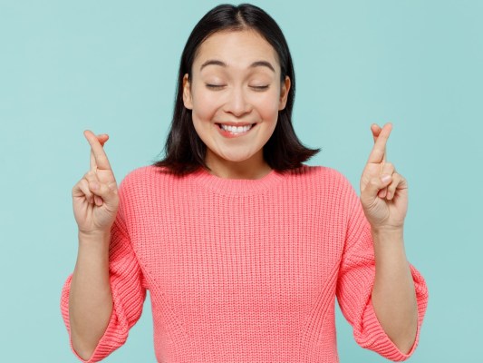 Woman of Asian descent in hot pink shirt, smiling with closed eyes and both sets of fingers crossed. She is posing against a light blue backdrop