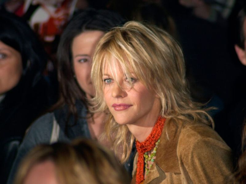 Meg Ryan in a crowd of people wearing a brown jacket and red scarf.